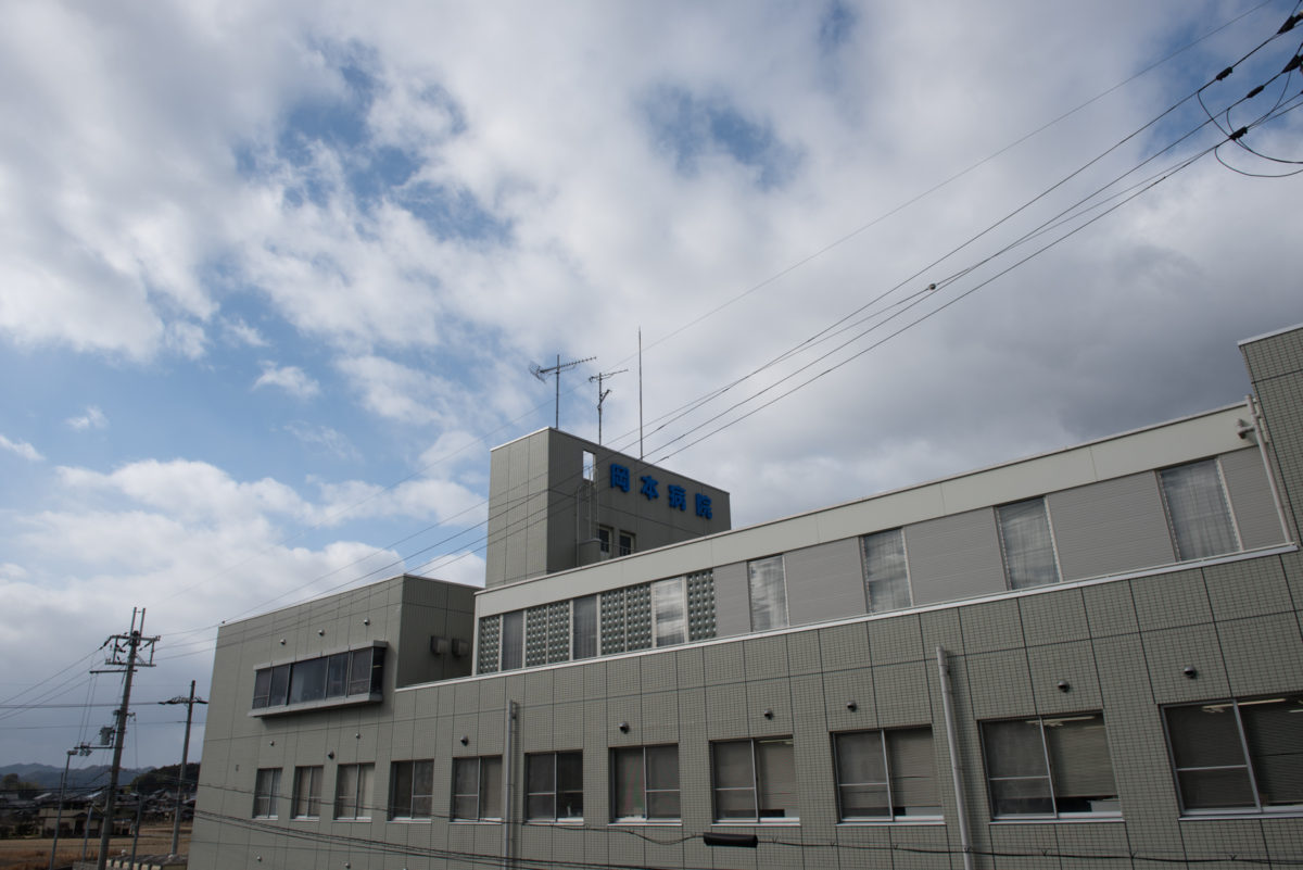 介護施設の建物外観。白色系の平屋造りで、上部に青い標識がある。